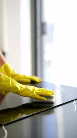 
A professional cleaner wearing yellow gloves scrubs the induction stove surface with a sponge, removing grease and stains during kitchen cleaning