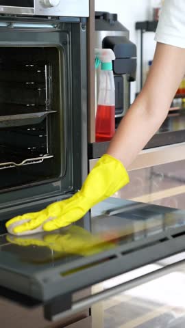 A professional cleaner wearing yellow gloves carefully wipes the oven door with a cloth and cleaning spray during kitchen deep cleaning
