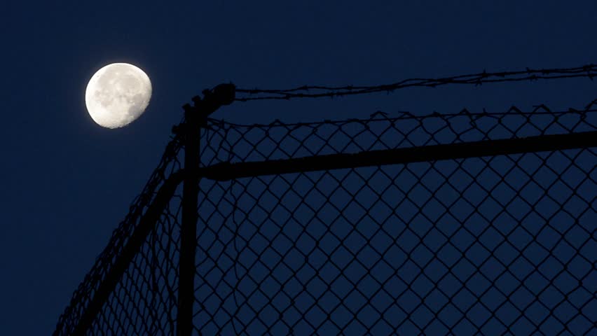 Time lapse, A bright full moon climbs above a barbed-wire fence and chain-link barrier, casting silhouettes in the dark sky. The image evokes solitude, boundary, and quiet nocturnal atmosphere.