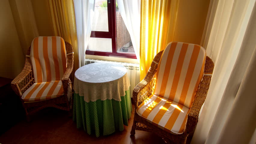 Warm seating nook featuring two wicker chairs with yellow and white striped cushions, a round lace-covered table, and bright sunlight streaming through sheer curtains by a window corner. Time lapse