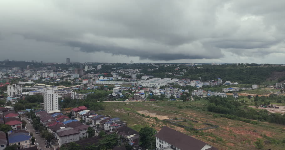 Zoomed out shot shows a heavy storm cloud looming over Sihanoukville, capturing a sprawling landscape that mixes urban development, green hills, and large plots of cleared land for construction.