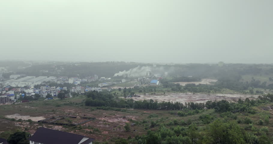 hazy view of a developing landscape, where a plume of white smoke rises from buildings scattered across green hills, with a muddy construction site in the foreground under a light rain