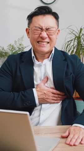 Stressed Asian businessman freelancer suffers from tension holding chest with heart pain attack risk of stroke symptom while working at modern home office workplace desk. Male Chinese in formal suit