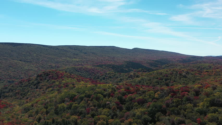 Seneca Creek Backcountry in West Virginia, with its rolling mountains covered in colorful red and green autumn foliage, is captured in an aerial shot moving forward under a blue sky.