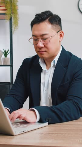 Tired Asian middle-aged businessman suffers from spine neck muscle pain at home office workplace working on laptop. Overworking. Freelancer Chinese man in suit attempt to relieve pain by massaging