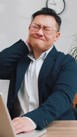 Tired Asian middle-aged businessman suffers from spine neck muscle pain at home office workplace working on laptop. Overworking. Freelancer Chinese man in suit attempt to relieve pain by massaging
