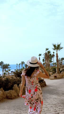 Smiling, beautiful young woman, in sunglasses, light summer dress, fluttering in wind, and sun hat, walking along street, with palm trees, by the sea. hot summer day