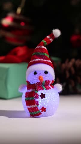 A small snowman figurine with a striped hat and scarf glows with different colors. A Christmas tree and wrapped gifts are in the background creating a holiday atmosphere.