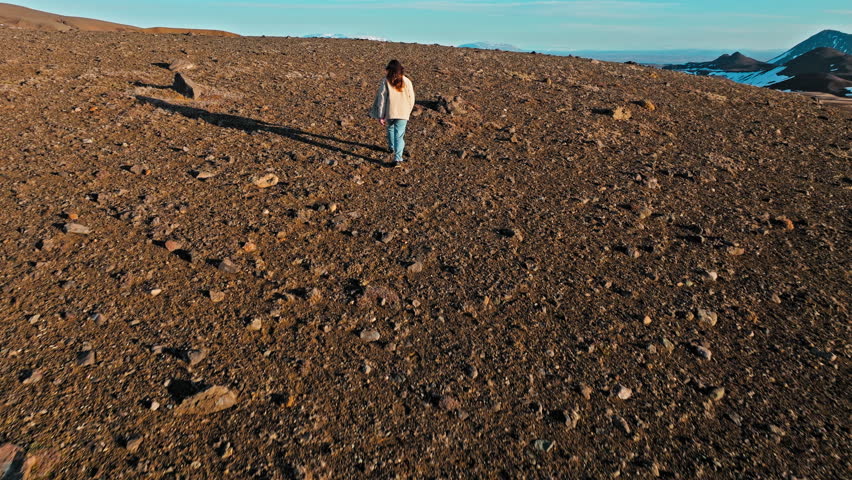 Female tourist walking across volcanic terrain at Krafla crater in North Iceland. Red slopes and melting snow contrast against deep blue water.