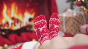 A person wearing red Christmas socks with reindeer patterns relaxes by a fireplace, surrounded by wrapped presents and a festive tree. The scene evokes warmth, comfort, and holiday spirit - Powered by Shutterstock - Get 15% off with code: PIKWIZARD15