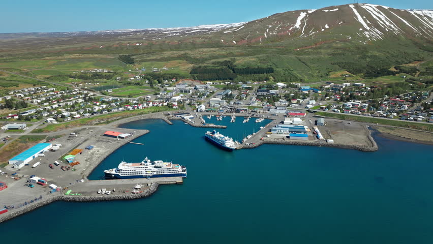 Drone shot of Husavik harbor with ships docked along the pier. The coastal town is surrounded by green hills and snowy peaks.