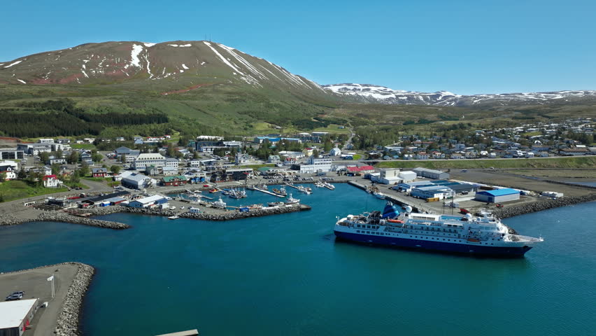 Drone view of Husavik harbor and cruise ship. The North Iceland port sits beside calm blue water and green mountain slopes.