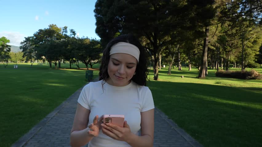 A smiling Colombian woman walks through a park on a sunny afternoon while searching for a location on her ph