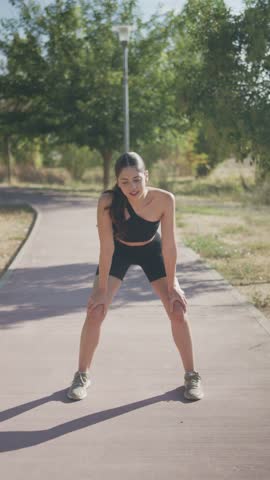 Tired fit woman taking a break from running in a park, smiling at the camera on a sunny day