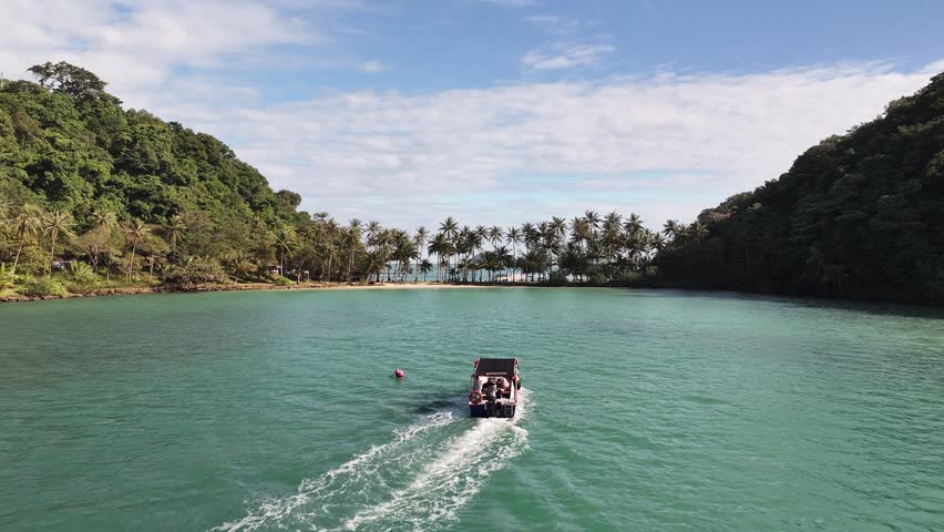 Cinematic 4K drone shot of a boat sailing toward a tropical palm beach with turquoise sea and jungle hills – plan contemplatif de voyage en bord de mer.
