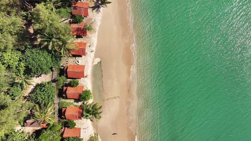 Cinematic 4K top-down drone shot of tropical beach huts with red roofs, turquoise water and palm trees – plan contemplatif aérien d’une plage tropicale paisible.