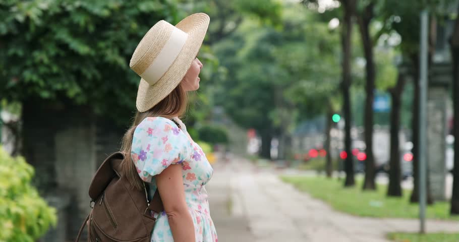 Woman in summer dress and hat photographing urban scenery with camera outdoors, exploring city street with trees, backpack on shoulder. Concept of travel, tourism, adventure.
