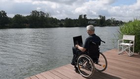 Elderly individual in wheelchair interacts with laptop at lakeside, highlighting the blend of technology and nature, promoting accessibility and outdoor engagement - Powered by Shutterstock - Get 15% off with code: PIKWIZARD15