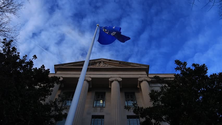 The Department of Justice (DOJ) flag waves in the wind, captured in a low-angle shot in front of the classical facade of its Washington D.C. headquarters, against a deep blue, cloudy sky.