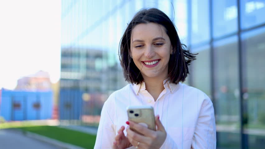Joyful female executive using her cellphone while strolling outside in a contemporary cityscape, with a sleek glass skyscraper in the background