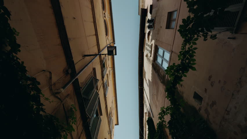 Narrow Roman street view from below with historic buildings, window shutters, lantern, and leafy plants framing the sky. High quality 4k footage