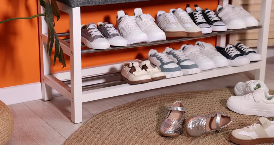 Different kid's shoes on storage bench and floor near orange wall indoors. Camera moving right