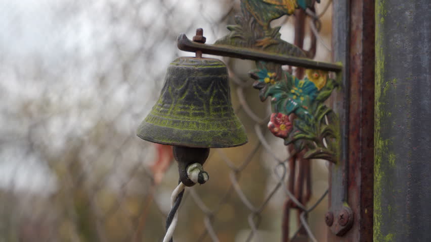 A rustic, dark metal bell with mossy green residue is hanging on a weathered, rusty gate post and attached to an ornate, painted metal bracket. It is outdoors with a blurred wire fence