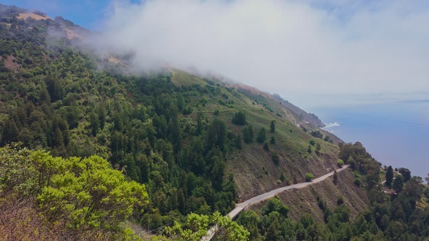 Video footage of Highway 1 winding through Big Sur, California, USA. Scenic coastal road with ocean cliffs, green forested hills, and drifting fog. Dynamic travel footage shot from a moving car