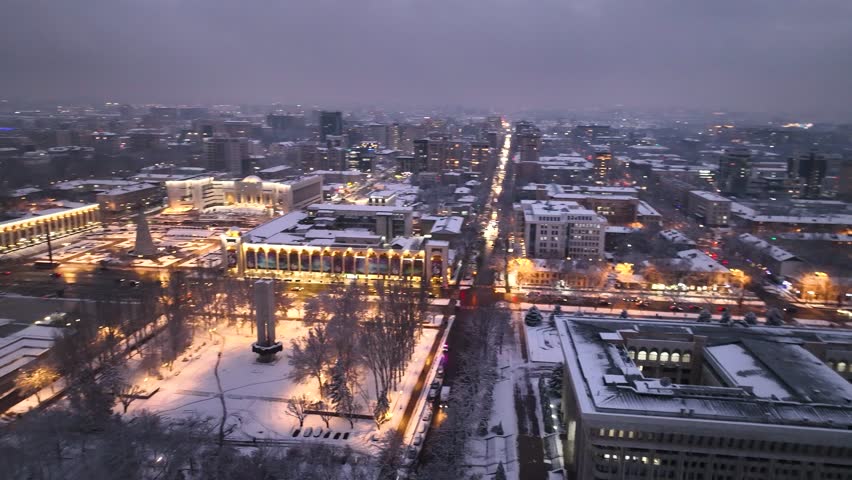 Wide aerial view of the snow-covered city center at twilight, dominated by the brightly illuminated Kyrgyz National Philharmonic Hall and surrounding urban grid