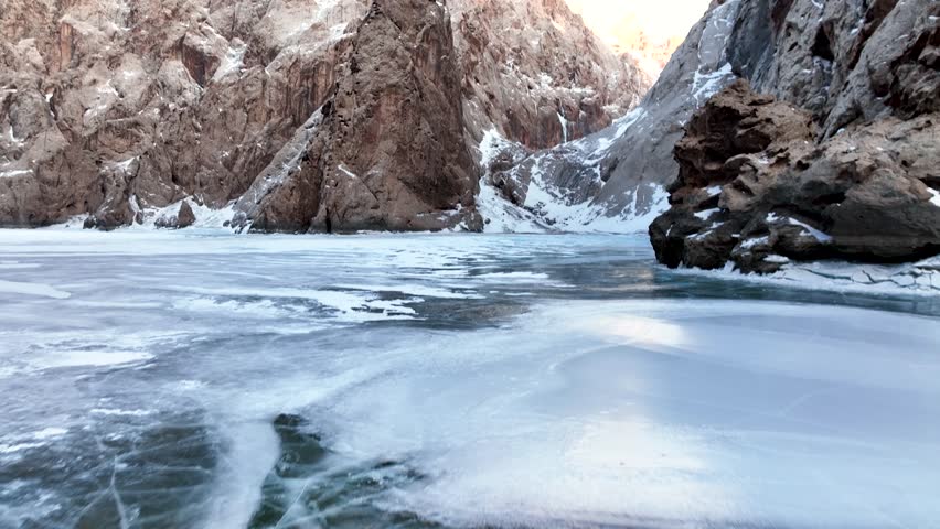 Low angle view from the surface of a frozen, cracked lake or river leading into a deep mountain canyon with towering, sheer rock walls dusted with snow and ice