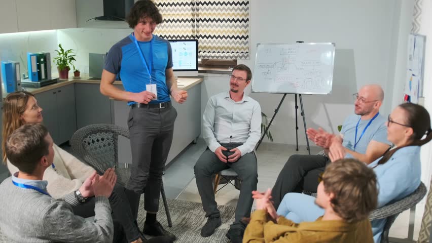 Young man in a blue T-shirt stands up at a psychotherapy meeting, showing his badge and introducing himself as part of a corporate training on trust and relationships. Colleagues applaud in support.