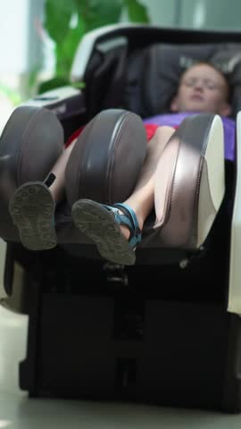 A teenager in a massage chair enjoys and relaxes from a machine massage in a public place. Portrait of a boy receiving a machine massage. 