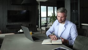 Young businessman sitting at a table at home working on a laptop and writing notes down on a notepad. - Powered by Shutterstock - Get 15% off with code: PIKWIZARD15