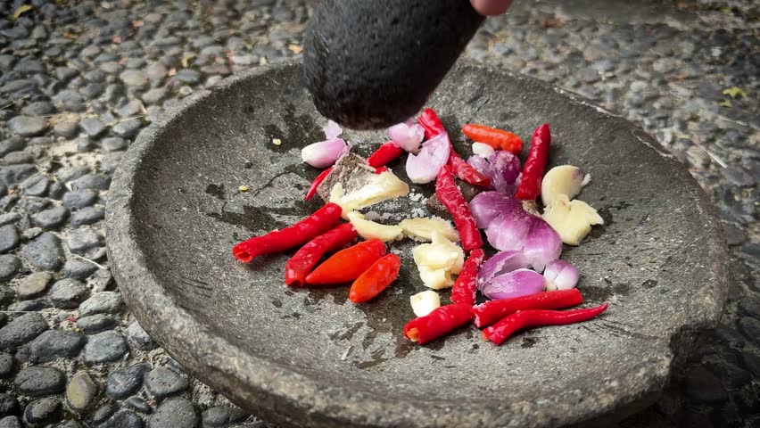 The making of chili paste called sambal , grinding red chili peppers, onion, and garlic in a rough stone mortar and pestle on a cobblestone surface, conveying rustic cooking and fresh spices, 