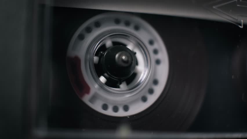 Macro view of a transparent audio cassette playing in a deck, with a white plastic reel spinning quickly. Dark magnetic tape unwinds, revealing a red leader tape section in the dark interior.