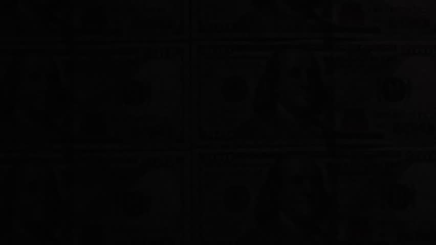 Overhead view of a hand holding a UV flashlight to check sheets of uncut hundred dollar bills. The purple beam scans across the currency, testing for authenticity and revealing hidden security marks.