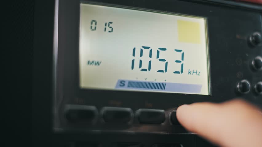 Macro of a finger cycling through FM SW and LW radio bands on a digital receiver. The backlit LCD screen displays changing frequencies a preset station number and a signal strength indicator.