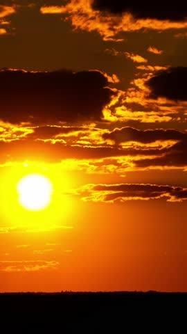 Vertical timelapse of golden sun setting behind dramatic fiery-edged clouds. Intense orange and red glow casts over a silhouetted horizon.