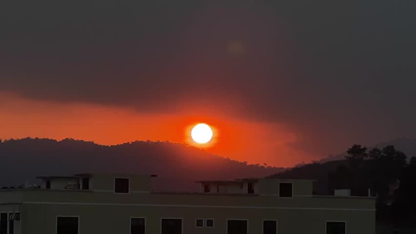 A vivid sunset in Titi New Village, Malaysia. The orange-red sun cuts through clouds and haze, lighting up the horizon, distant hills, and building rooftops with dramatic atmospheric color.