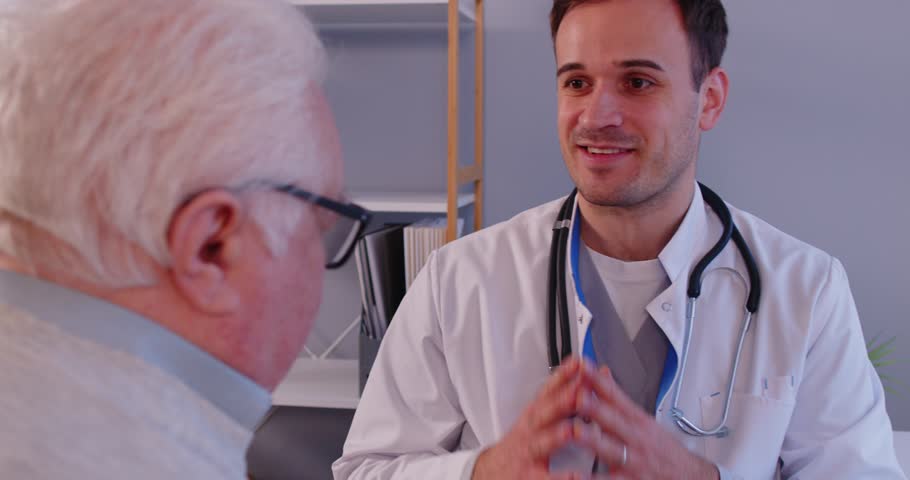 Positive male doctor or nurse provides consultation and emotional support to an elderly patient in a hospital office. Medical care, compassion, and professional assistance for senior people.