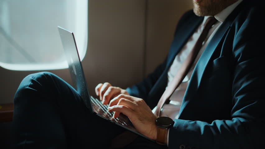 Productive middle-aged businessman in a formal suit typing on a laptop keyboard while traveling on an airplane, sitting comfortably in a business class seat next to the window