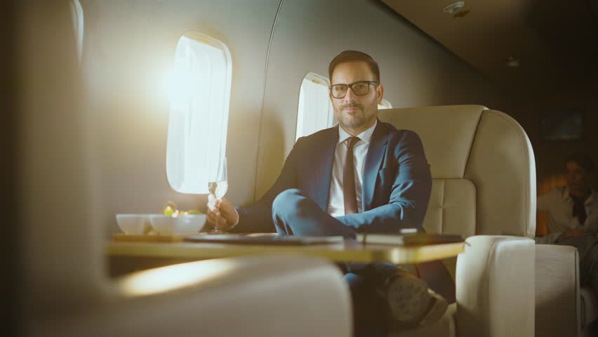 Handsome successful businessman in a suit drinking champagne and smiling at the camera while traveling in first class on a private jet, enjoying a luxurious lifestyle and business trip