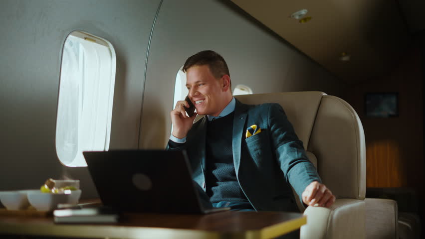 Confident businessman traveling on a private jet, enjoying a phone conversation and smiling while sitting in a comfortable leather seat with a laptop on the table next to him