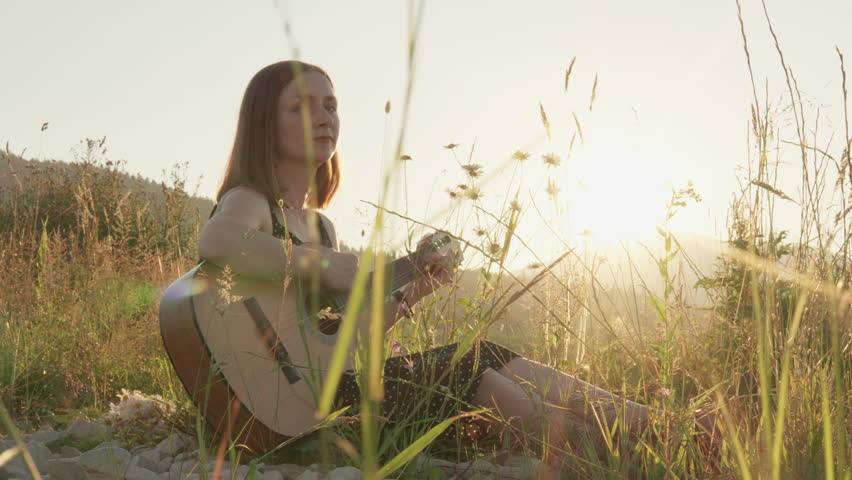 A young woman sits serenely in a lush grassy field, skillfully playing her acoustic guitar as the sun sets, casting a warm and golden light that beautifully envelops the idyllic landscape around her
