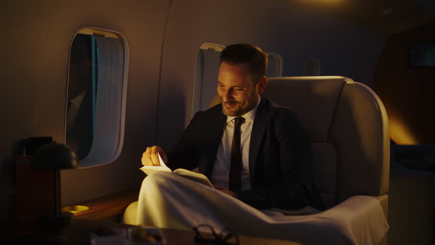 Happy millionaire in a formal suit relaxing while reading a book during a comfortable night flight. Luxurious business travel experience inside a modern private jet cabin for a first-class journey
