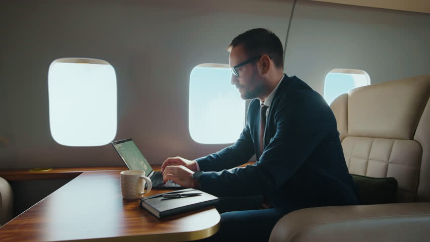 Confident businessman in a suit working on a laptop while flying in a luxurious private jet. Successful executive managing his business during a comfortable and exclusive first-class flight
