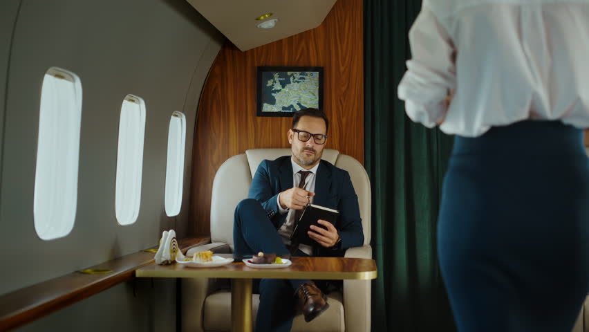 Professional flight attendant serving champagne to a successful businessman traveling in a luxurious private jet, while the passenger is taking notes on his personal agenda on his way to work
