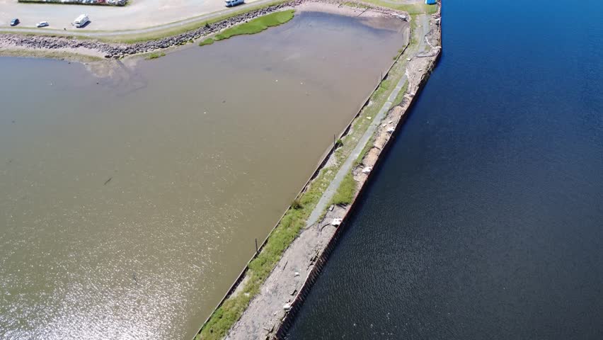 Aerial view of the remains of an old abandoned wharf separating two bodies of water of different colors, blue and brown, with the reflection of the sun sparkling on the waves. Campbellton, NB, Canada.