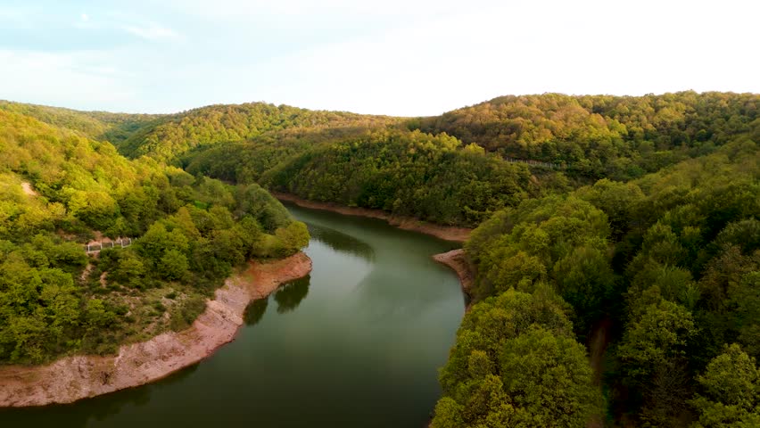 Aerial view of a winding river surrounded by forested hills during golden hour. Scenic nature landscape with lush trees, erosion textures, and tranquil water. Ideal for drone, forest, and reservoir th