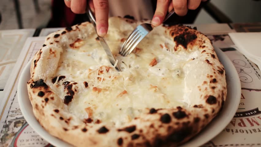 Close up of a man cutting hot cheese pizza with a fork and knife in an outdoor cafe. Warm tones and delicious melted cheese. Horizontal 4k footage
